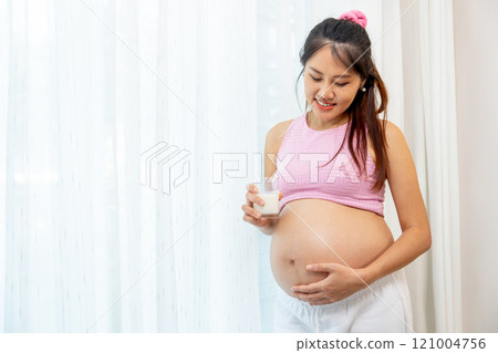 Asian pregnant woman stand with hold a glass of milk and stand in front of white curtain also look at her belly with happiness smile. Asian pregnant woman stand with hold a glass of milk and stand in front of white curtain also look at her belly with happiness smile. 121004756