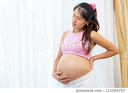 Asian pregnant woman stand in front of white curtain and action like she feel backpain that she touch her back and belly. Asian pregnant woman stand in front of white curtain and action like she feel backpain that she touch her back and belly. 121004757