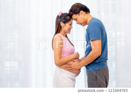 Asian man and pregnant woman stand with hug and attach forehead together express love and care in front of white curtain. 121004765