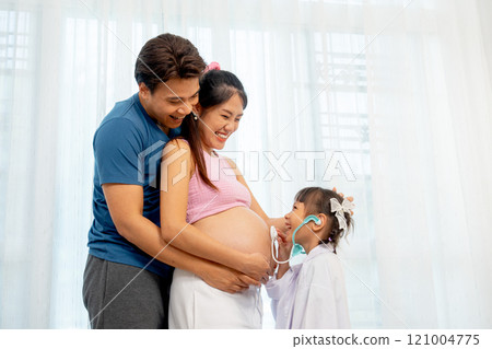 Asian father hug pregnant mother in her back and little girl use stethoscope toy to play with belly of mother in front of white curtain in their house. 121004775