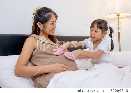 Wide shot little Asian girl enjoy to play with pink baby shoes on belly of pregnant mother and stay on bed of their house to express love and care in family. 121004840