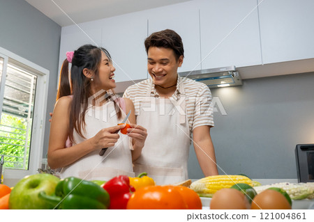 Asian pregnant woman use knife to prepare or cook for man who stand beside in the kitchen of their house and they also look to each other with happiness. 121004861