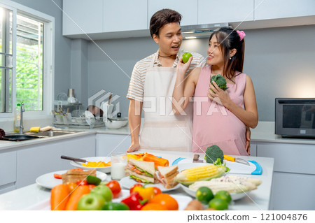 Asian man stand beside pregnant woman who hold apple and vegetable and give to man for eating in the kitchen of their house. 121004876