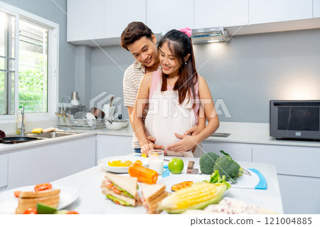 Asian man stand and hug from the back of pregnant woman and during woman prepare to cook in the kitchen of their house. Asian man stand and hug from the back of pregnant woman and during woman prepare to cook in the kitchen of their house. 121004885