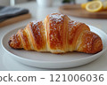 Golden brown croissant with flaky layers, dusted with powdered sugar, served on white plate. Perfect for breakfast or delightful snack 121006036