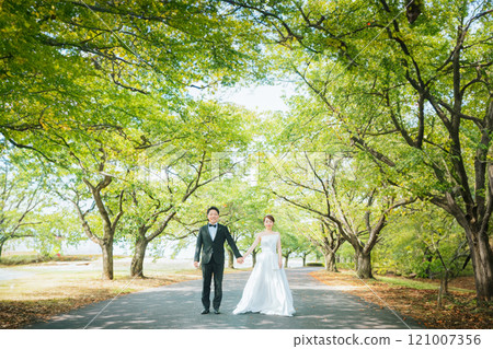 一對穿著禮服的情侶手牽手沿著公園的綠樹成蔭的大道散步（婚禮、婚紗照、新娘和新郎） 121007356