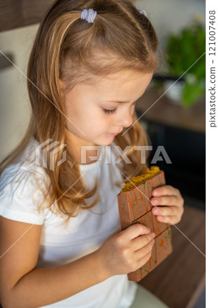 Little girl eating Dubai chocolate with pistachio paste and kataifi dough. Confectionery handmade sweets at home in the kitchen.  121007408