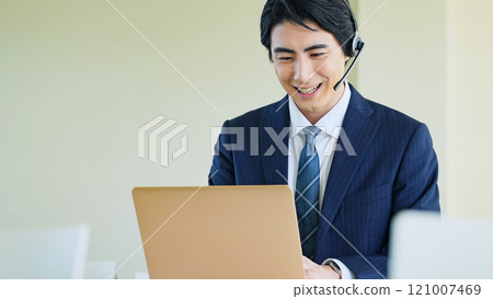Male business person talking on a headset while looking at a laptop 121007469