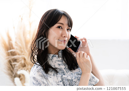 A young woman with long black hair sitting on a white sofa and talking on her smartphone 121007555