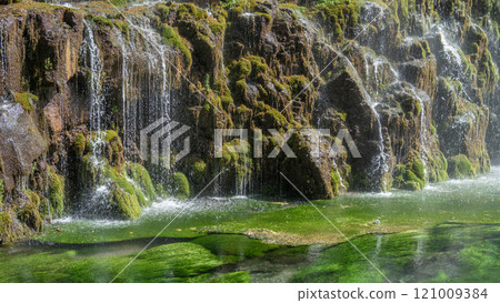 A serene waterfall cascades over moss-covered rocks into a clear, green mountain river. A serene waterfall cascades over moss-covered rocks into a clear, green mountain river. 121009384