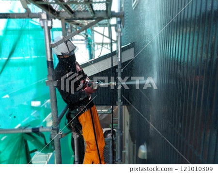A male craftsman uses high-pressure washing to prepare the exterior walls of a wooden house for painting 121010309