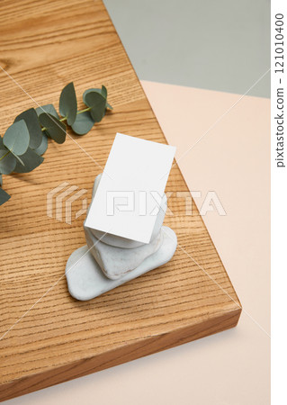 Blank card standing upright against stacked white stones on wooden block, complemented by eucalyptus sprig against soft tones. Blank card standing upright against stacked white stones on wooden block, complemented by eucalyptus sprig against soft tones. 121010400