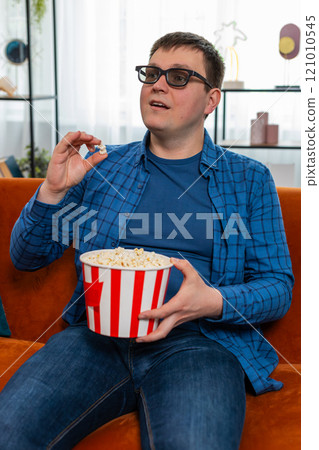 Caucasian man in 3D glasses sitting on couch eating popcorn snacks and watching interesting TV film 121010545
