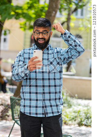 Indian man use mobile smartphone celebrating win good message news outdoors on urban city street 121010573