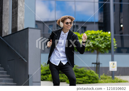 Happy relaxed young Caucasian businessman in wireless headphones dancing, listening music outdoors 121010602
