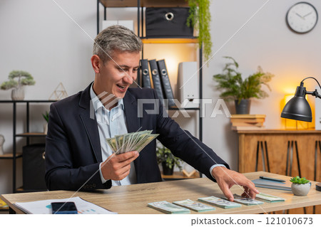 Happy young Caucasian successful businessman in formal suit counting money cash at home office desk 121010673