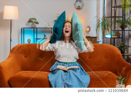 Irritated young woman reading book cover ears with pillows annoyed by loud noise sitting on sofa 121010684