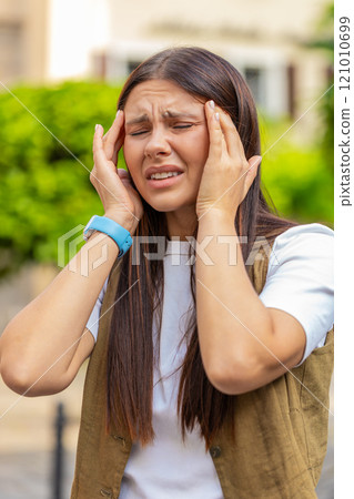 Displeased exhausted young Caucasian woman rubbing temples to cure headache standing on city street 121010699