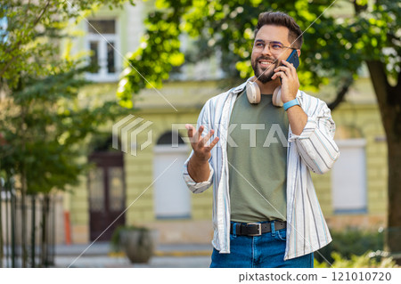 Happy young Caucasian man tourist in casual talking on smartphone while standing on city street 121010720