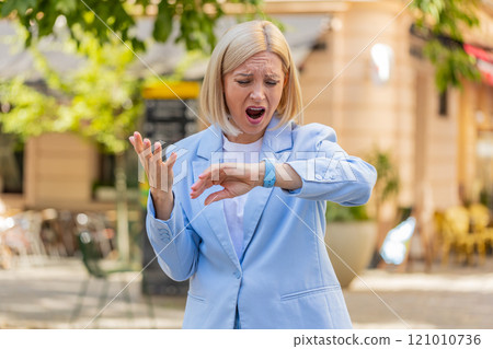 Shocked mature woman businesswoman in hurry checking time on smartwatch walking on city street Shocked mature woman businesswoman in hurry checking time on smartwatch walking on city street 121010736