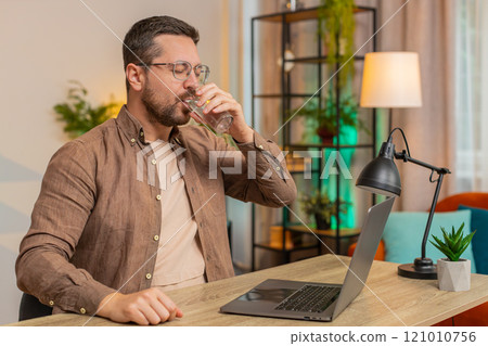 Focused Caucasian businessman sitting at desk drinking water while working on laptop in home office 121010756