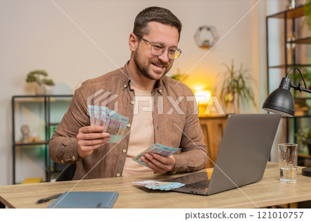 Rich happy Caucasian man counting money hryvnias cash using laptop calculate domestic bills at home 121010757