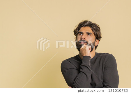 Thoughtful young Hispanic man looking aside with positive expression against beige studio background Thoughtful young Hispanic man looking aside with positive expression against beige studio background 121010783