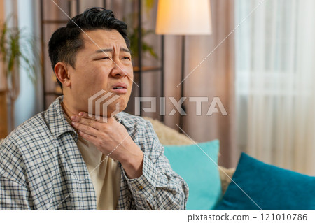 Upset Chinese young man holds hands over sore throat with painful neck and frowning on sofa at home Upset Chinese young man holds hands over sore throat with painful neck and frowning on sofa at home 121010786