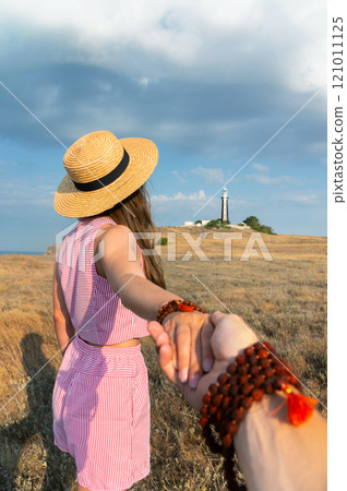 First person view of a man's hand holding the hand of his young companion while she looks into the distance at a landscape with a field and a lighthouse 121011125