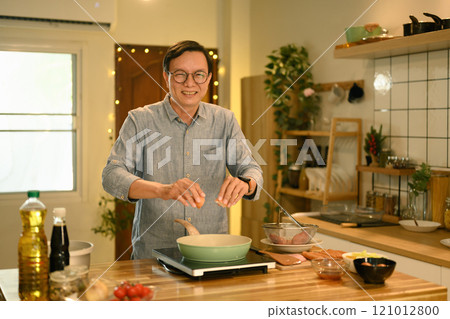 Cheerful middle aged man cracking an egg into a pan while cooking in a kitchen Cheerful middle aged man cracking an egg into a pan while cooking in a kitchen 121012800