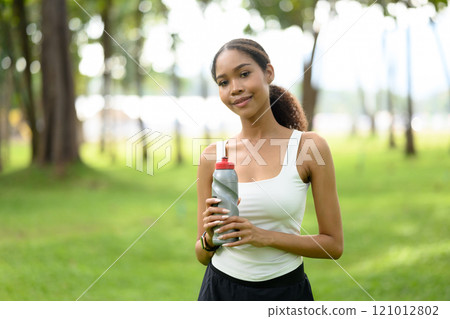 Young woman holding a water bottle while enjoying break during her outdoor exercise in a green park Young woman holding a water bottle while enjoying break during her outdoor exercise in a green park 121012802