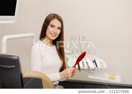 Pretty girl smiling while sitting in the dentist's chair, holding a mirror in her hands 121017890