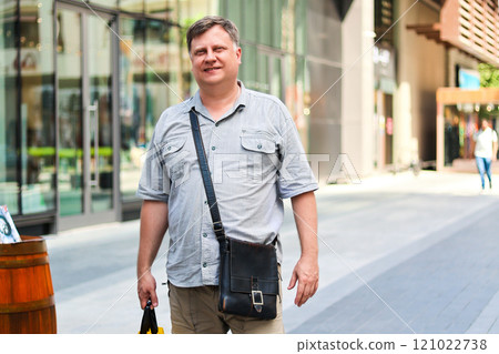 A Thoughtful Adult Man Carrying a Large Yellow Shopping Bag Filled with Groceries, Strolling Down the Street with a Content Smile, Embracing the Joy of Everyday Life and Weekend Errands 121022738