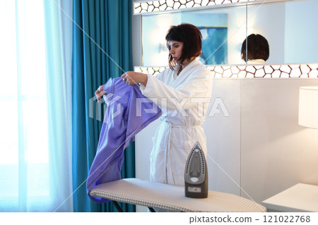 A thoughtful woman in a white dressing gown looks at her husband's things, irons them, enjoying the comfort of the home and the intimacy of their life together A thoughtful woman in a white dressing gown looks at her husband's things, irons them, enjoying the comfort of the home and the intimacy of their life together 121022768