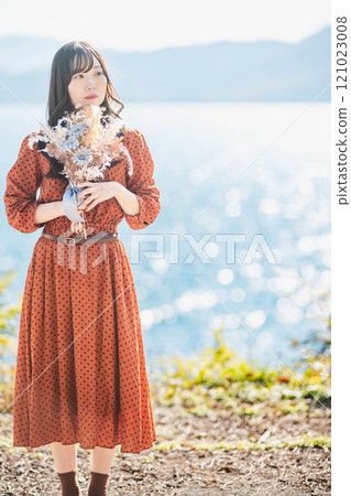 A woman in a red dress standing by a lake with dried flowers A woman in a red dress standing by a lake with dried flowers 121023008