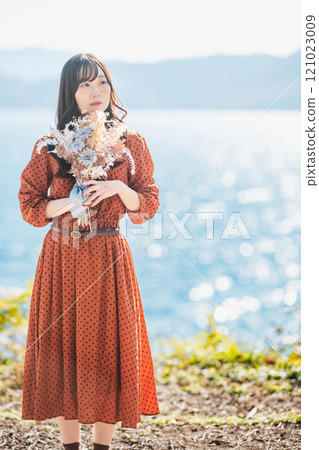一個穿紅裙子的女人站在湖邊，手裡拿著乾燥花 121023009