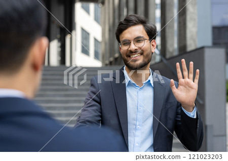 Businessman in suit greeting colleague with friendly smile and wave. Outdoor meeting in city, showcasing professional interaction and positive connection during workday. 121023203