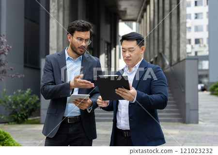 Businessmen discussing work in corporate environment. One holding tablet, another with clipboard, focused on conversation. Modern setting enhances business strategy context. 121023232