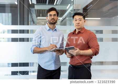 Two confident business professionals stand in a modern office setting, one holding a digital tablet and the other with a notepad. They exude focus and determination, emphasizing teamwork 121023390