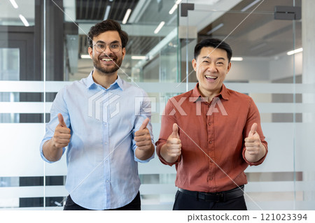 Two smiling men in an office setting, expressing positivity with thumbs up gestures, symbolizing success and teamwork. The image conveys a friendly and professional atmosphere 121023394