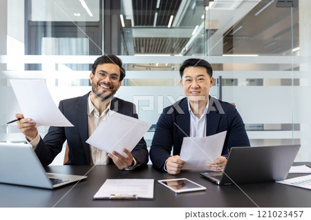 Smiling businessmen seated at office desk holding documents, highlighting teamwork and success. Laptops and paperwork reflect modern professional setting and cooperation. 121023457