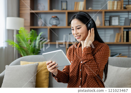 Asian woman at home enjoying music on headphones while using a tablet, seated on sofa in cozy living room. Scene depicts relaxation, technology, and casual leisure. Asian woman at home enjoying music on headphones while using a tablet, seated on sofa in cozy living room. Scene depicts relaxation, technology, and casual leisure. 121023467
