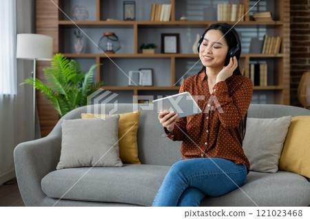 Asian woman enjoying music on headphones at home, sitting on comfortable sofa with tablet. Relaxation, leisure, technology. Cozy and peaceful setting indoors. 121023468
