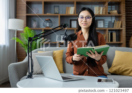 Asian woman recording podcast at home, using microphone, laptop, and book for engaging content creation. Casual setting enhances creative storytelling and personal connection. 121023497