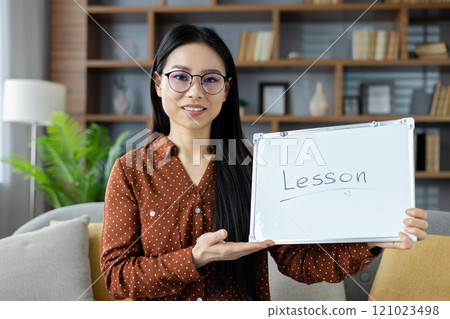Confident Asian woman holding whiteboard with 'Lesson' text, ready for online teaching or virtual training session in comfortable home setting. Wearing eyeglasses and professional attire. 121023498