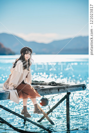 坐在湖邊的貝雷帽和風衣的年輕女子 坐在湖邊的貝雷帽和風衣的年輕女子 121023817