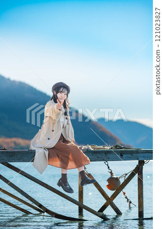 Young woman in beret and trench coat sitting by a lake 121023827