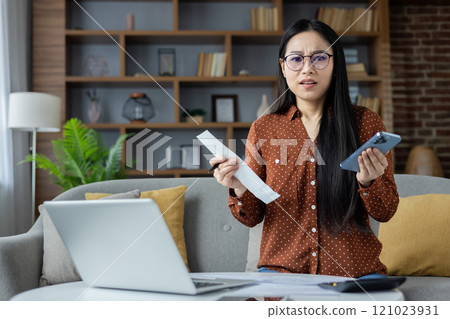 Asian woman holding receipt, phone while managing expenses in home setting. Expressing concern over financial issues with laptop on table. Business, finance, communication. 121023931