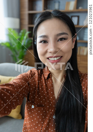 Asian woman captured in delight taking selfie with phone at home, seated on sofa, enjoying leisure time. Bright smile reflects happiness and comfort, framed by cozy living room interior. 121023961