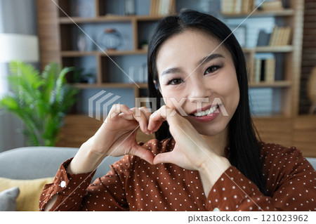 Happy Asian woman creating heart shape with hands, embodying love and joy. Smiling face and cozy home setting suggest warmth, affection, and positive energy, capturing a moment of heartfelt happiness Happy Asian woman creating heart shape with hands, embodying love and joy. Smiling face and cozy home setting suggest warmth, affection, and positive energy, capturing a moment of heartfelt happiness 121023962